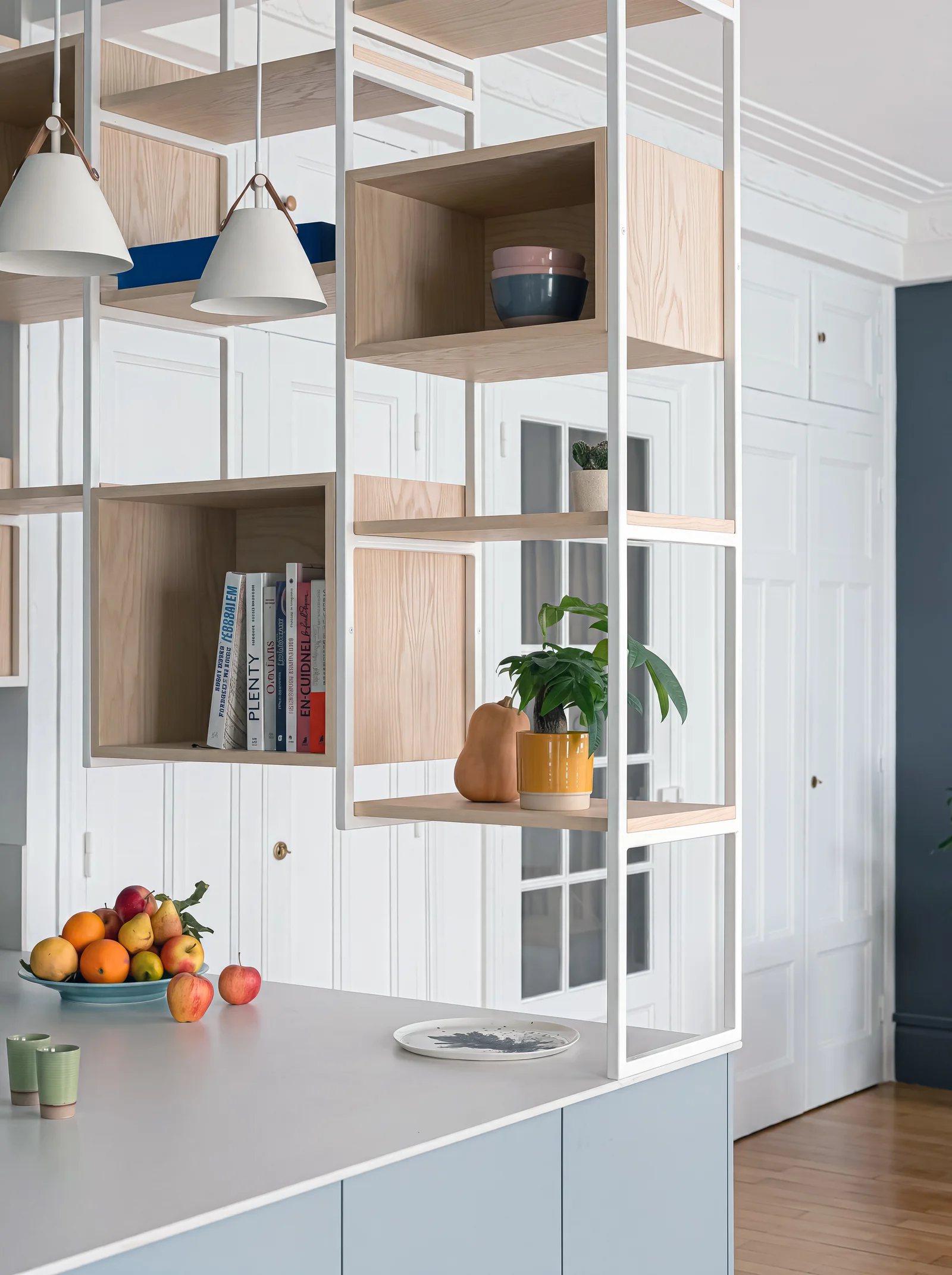 Kitchen peninsula with hanging white steel shelves and oak cubbies holding books, plants and fruit