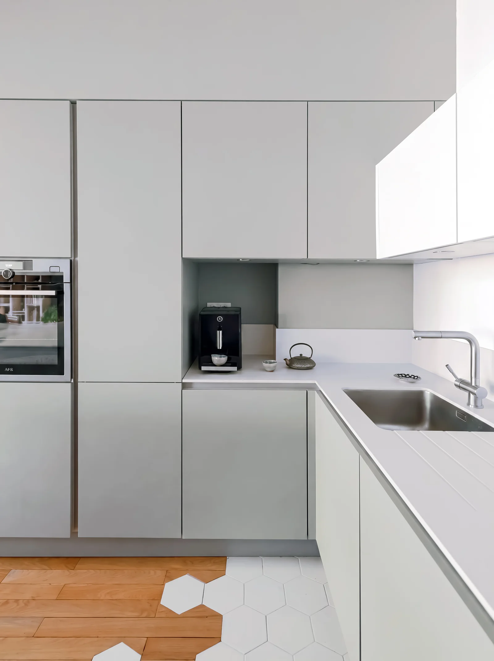 Corner of a grey handleless kitchen with tall units, a built-in oven and a pale worktop with sink