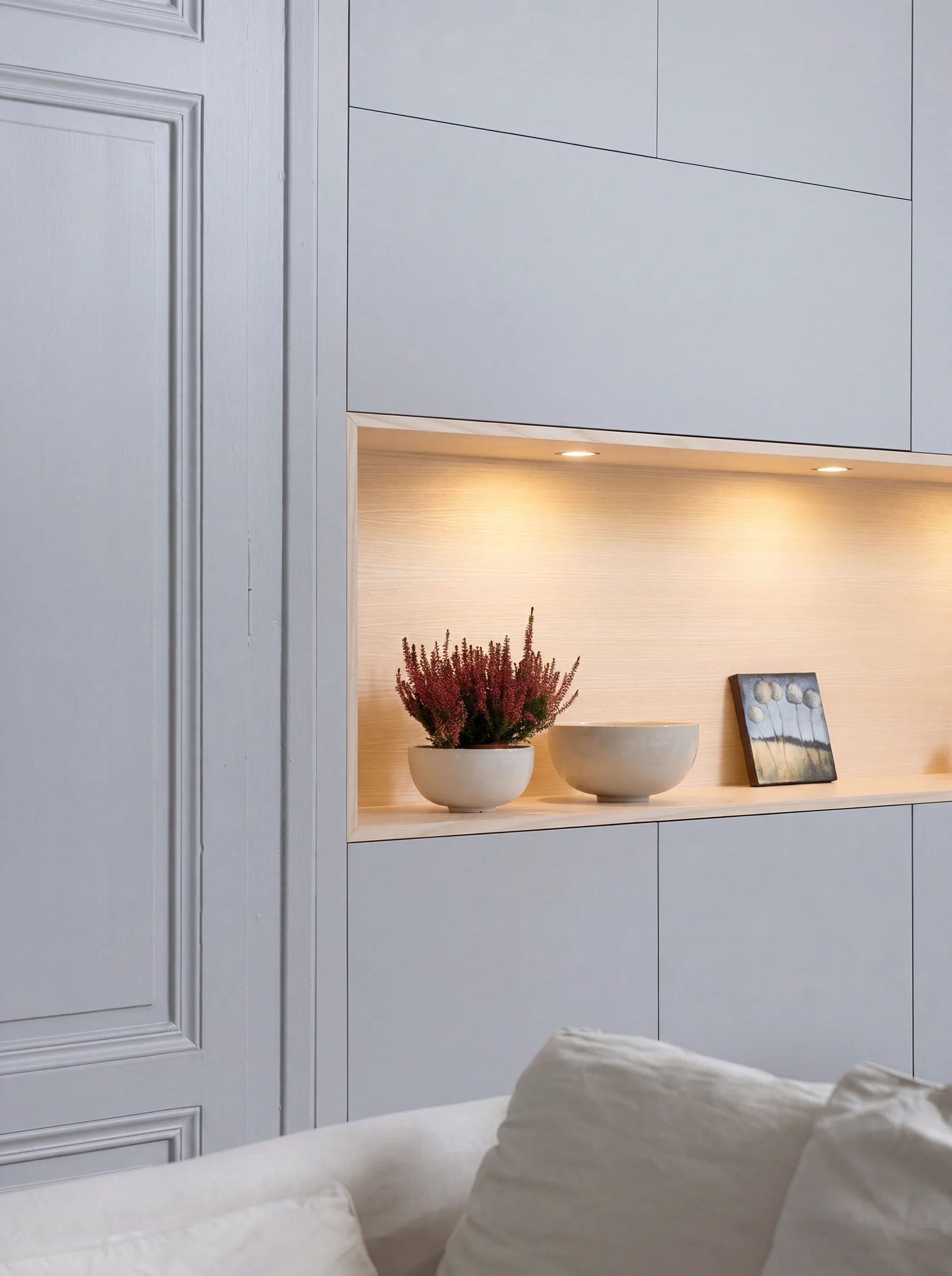 Close-up of a lit oak-lined shelf recess in pale grey panelled cabinetry with ceramics and heather