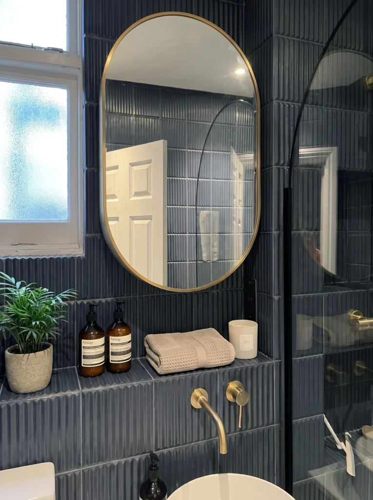 Navy tiled bathroom with a brass-framed oval mirror, a tiled ledge and a wall-mounted brass tap
