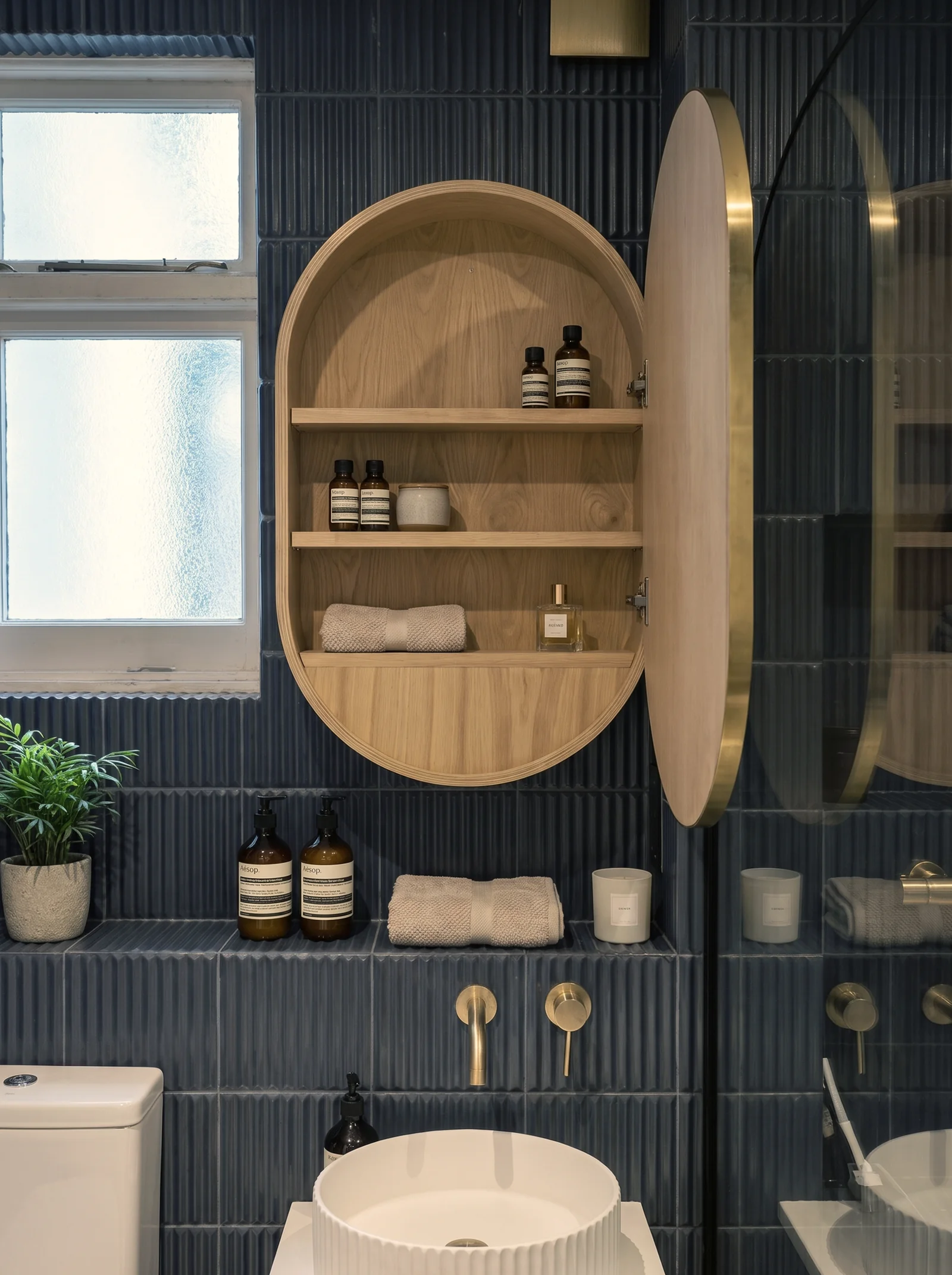 Oval oak bathroom cabinet with open shelves set into navy vertical tiles above a ribbed basin