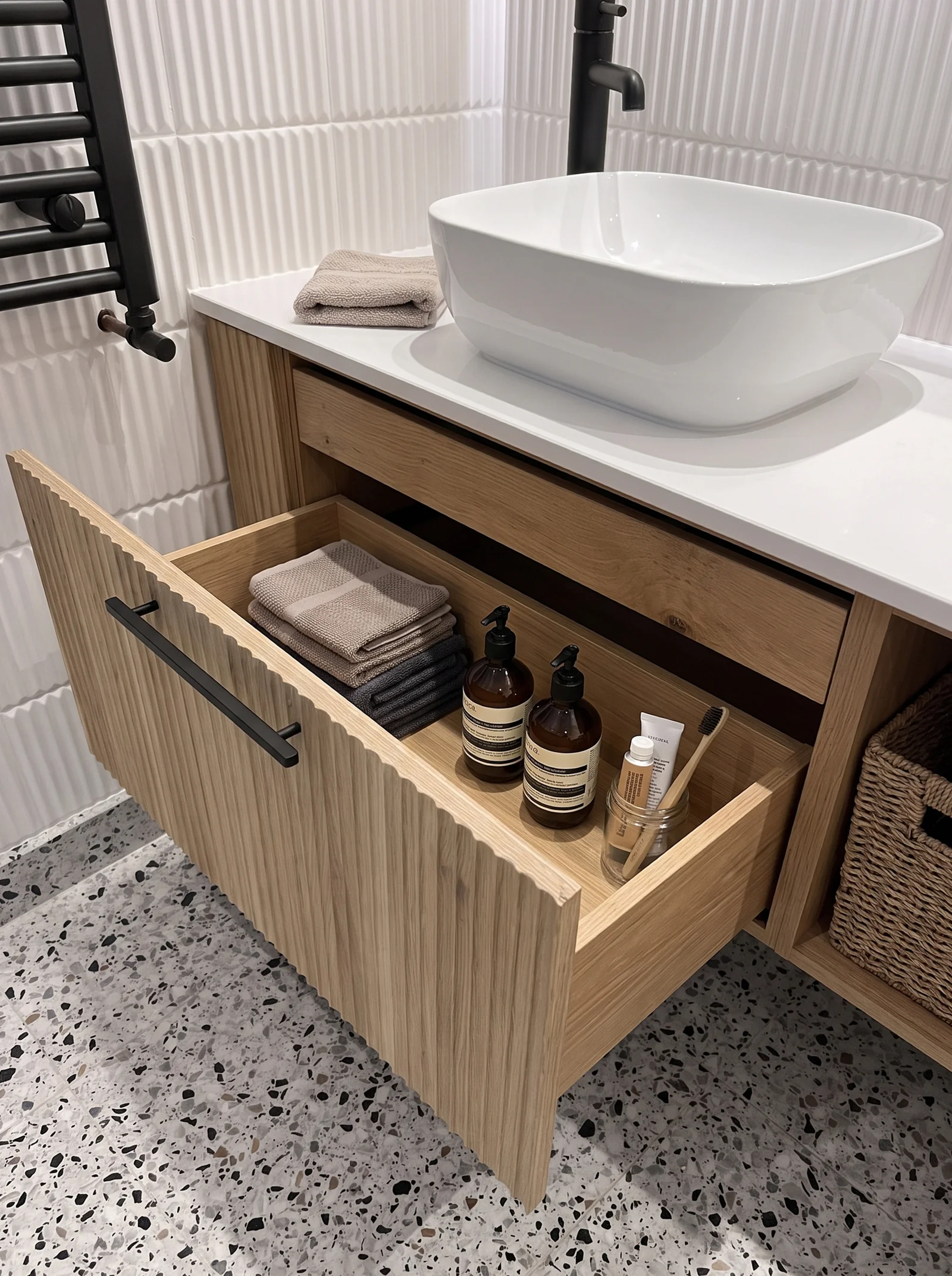Open oak vanity drawer holding towels and toiletries beneath a white vessel basin on a terrazzo floor