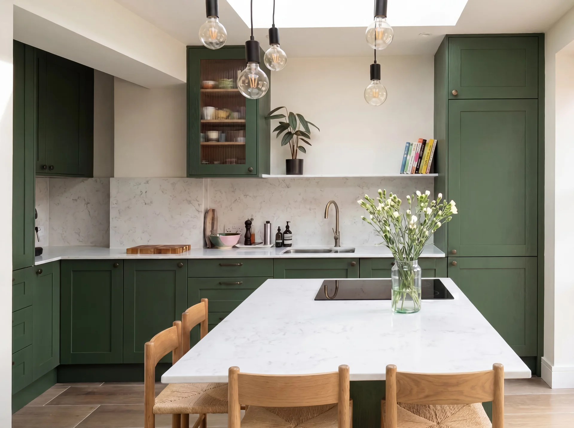 Deep green shaker kitchen with a glazed wall cabinet, white island worktop and oak stools under a skylight