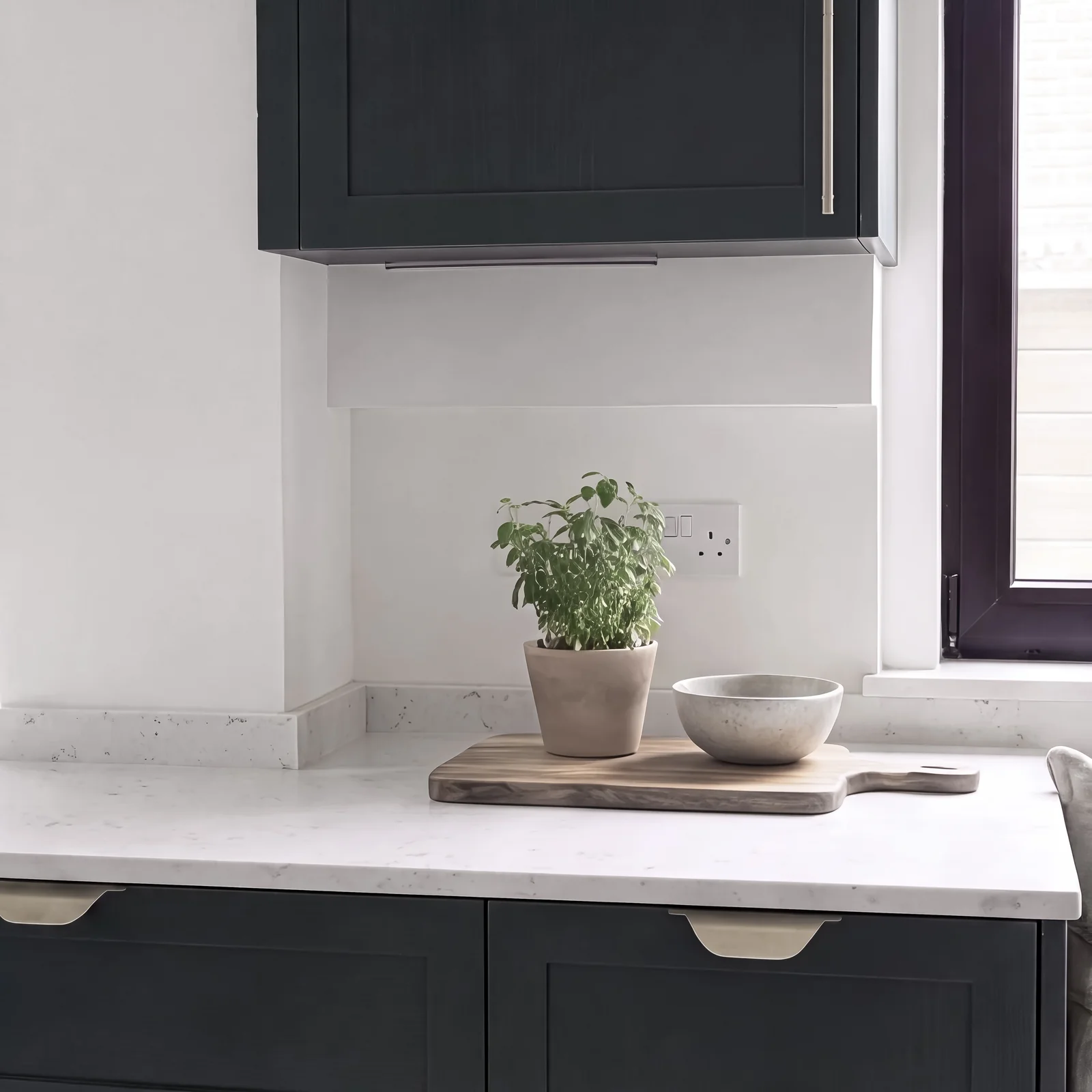 Potted herb and chopping board on a white worktop beneath a dark green wall cabinet