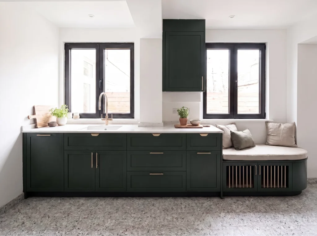 Dark green shaker kitchen under two windows, with a cushioned window seat over a slatted dog bed