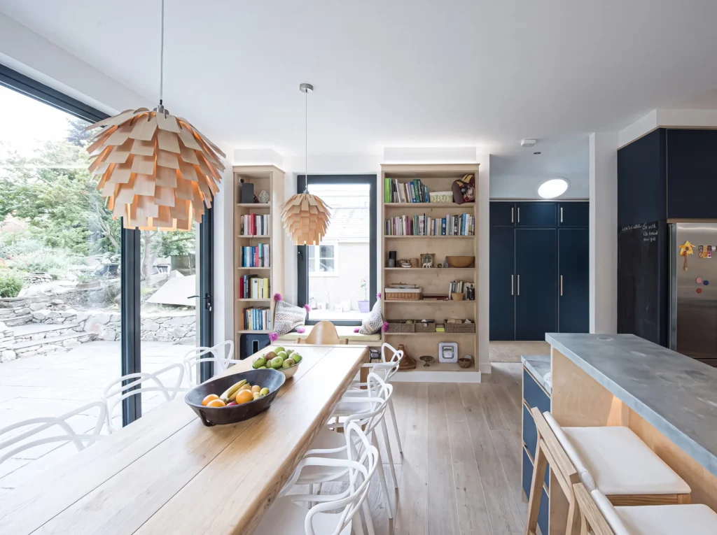Long open-plan kitchen with an oak dining bench, navy units and birch ply bookshelves by a garden window