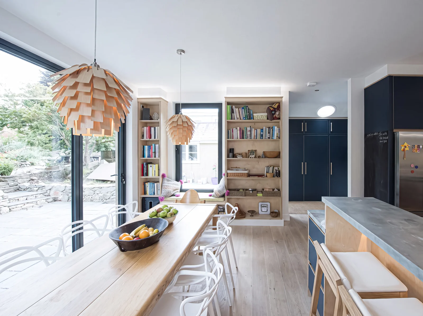 Long open-plan kitchen with an oak dining bench, navy units and birch ply bookshelves by a garden window