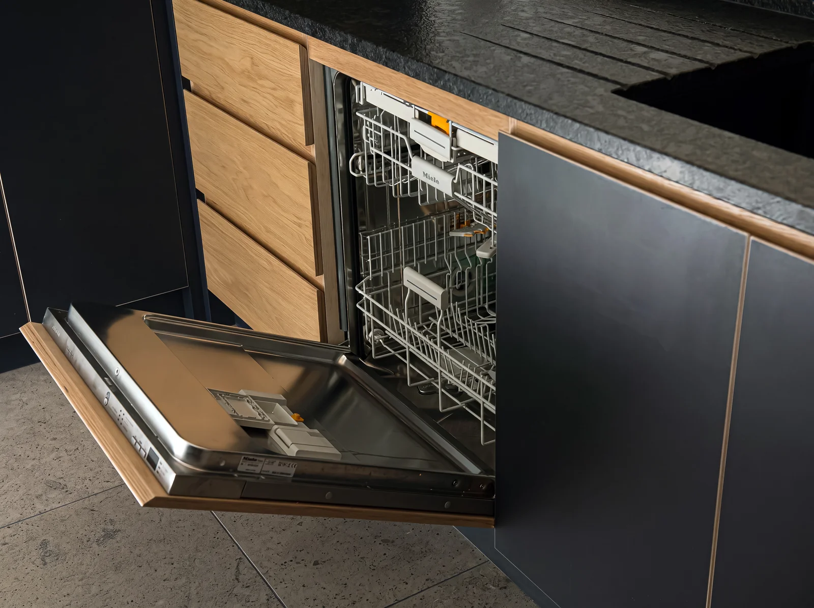 Integrated dishwasher open beneath a navy unit with oak drawers and a granite worktop