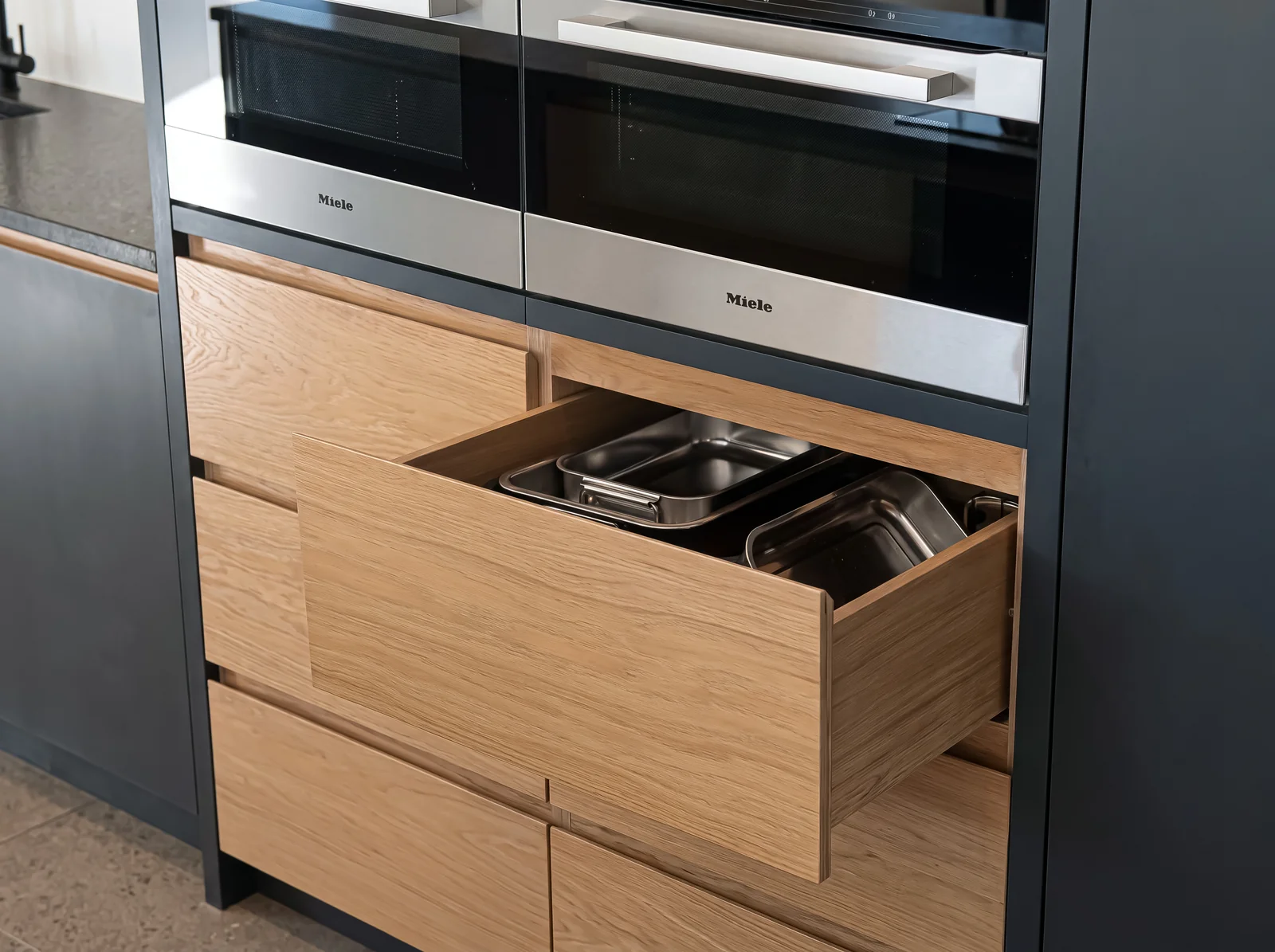 Open oak pan drawer beneath built-in ovens in a navy kitchen