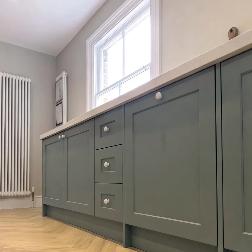 Run of darker sage shaker base units under a sash window beside a column radiator