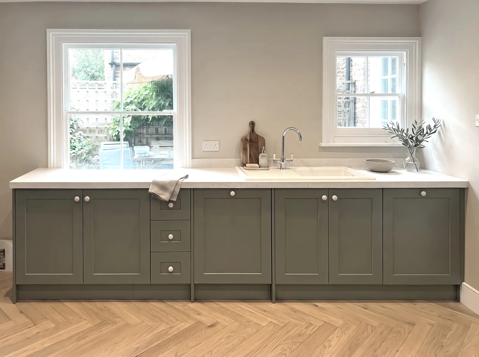 Sage shaker sink run with knob handles beneath two windows on a herringbone floor
