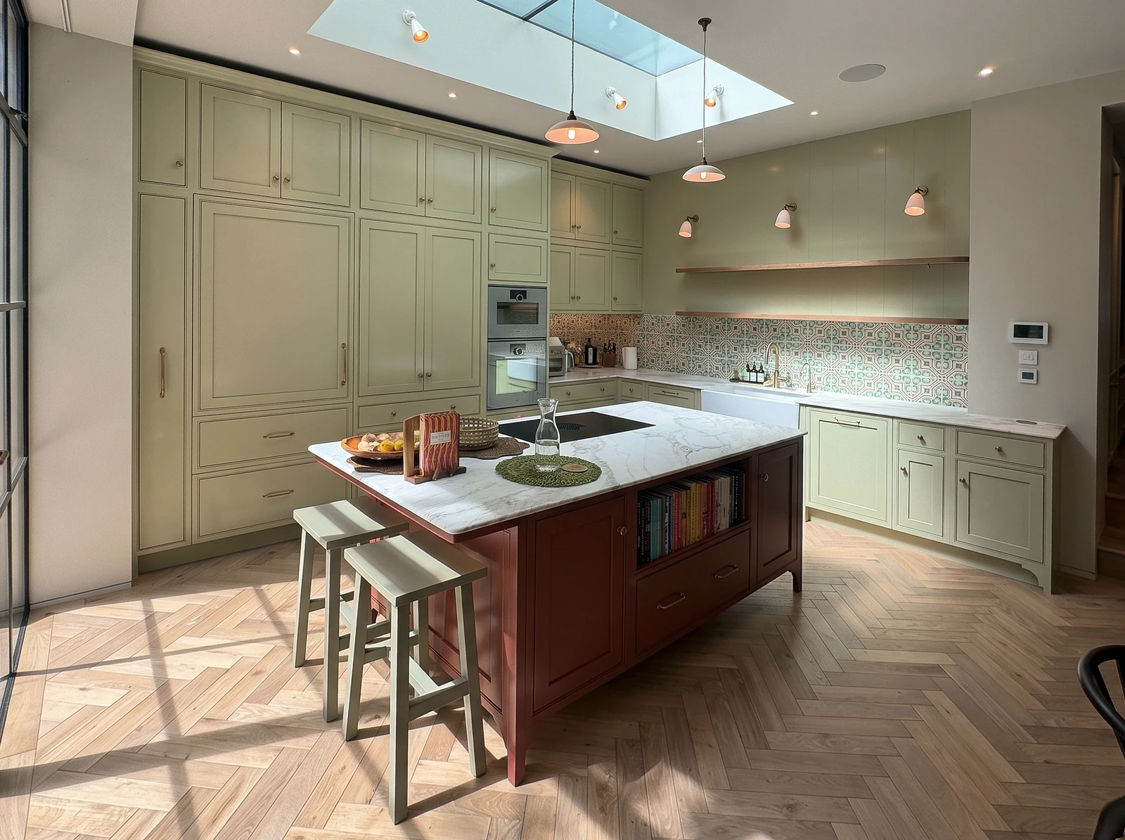 Sunlit sage kitchen with a deep red island, oak stools and a herringbone floor