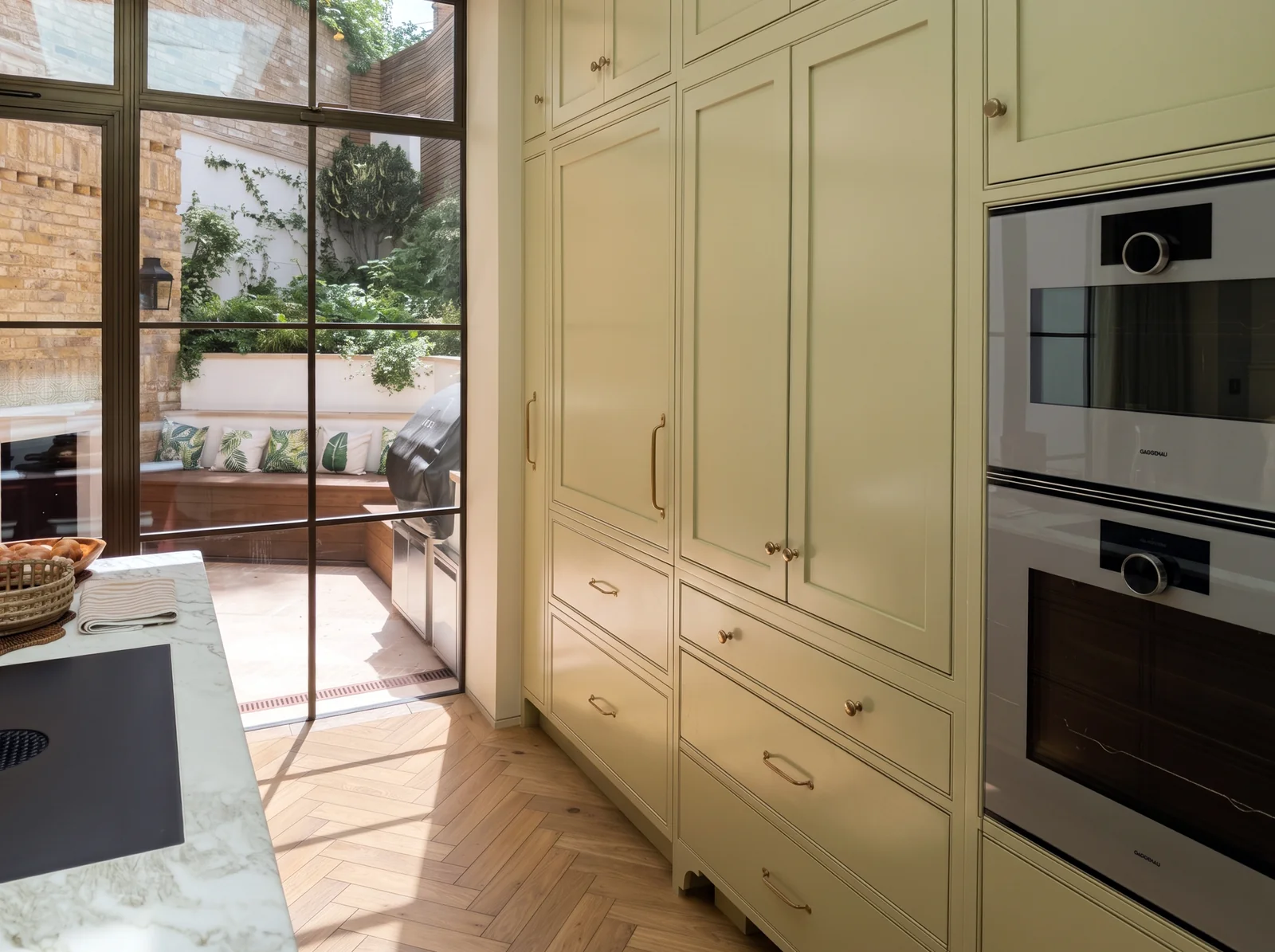 Tall sage shaker cupboards and double ovens beside black-framed garden doors