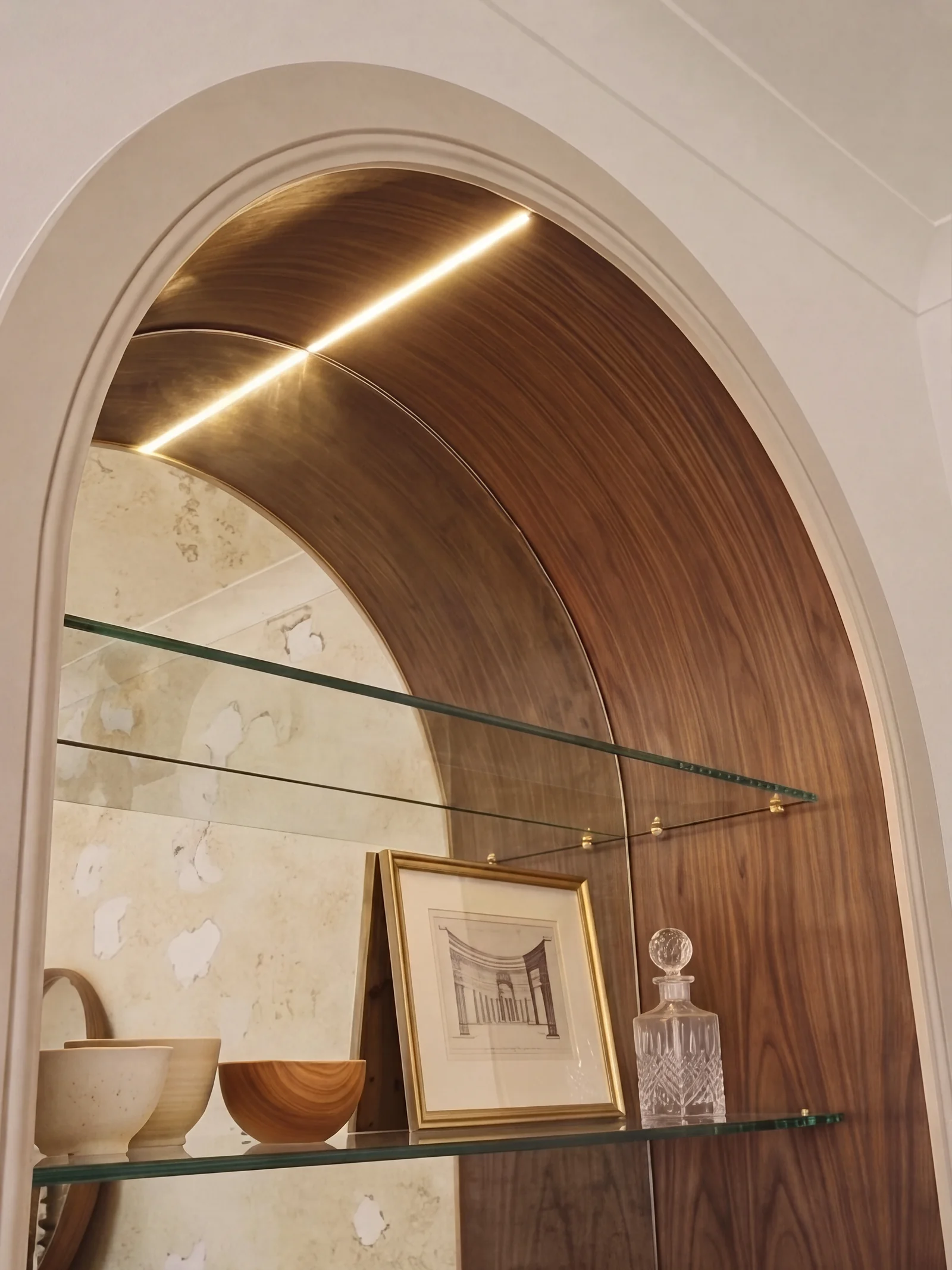 Lit arched walnut alcove with glass shelves holding a framed print and ceramics